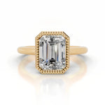 alt="An Emarald cut Moissanite engagement ring round shank with a bezel setting, on a gold band, against a white background.Moissanite engagement ring cape town south africa grand diamonds ralph jacobs jeulia 